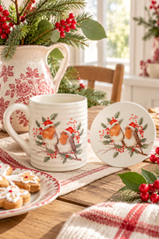 Tazza natalizia bianca in ceramica con due pettirossi e sottobicchiere coordinato, ambientati su tavolo rustico in cucina cottage.